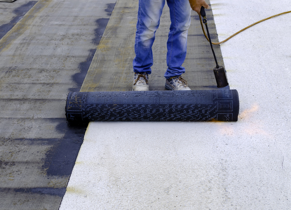 Person applying waterproofing membrane with a blowtorch to a flat roof. They are wearing jeans and work boots.