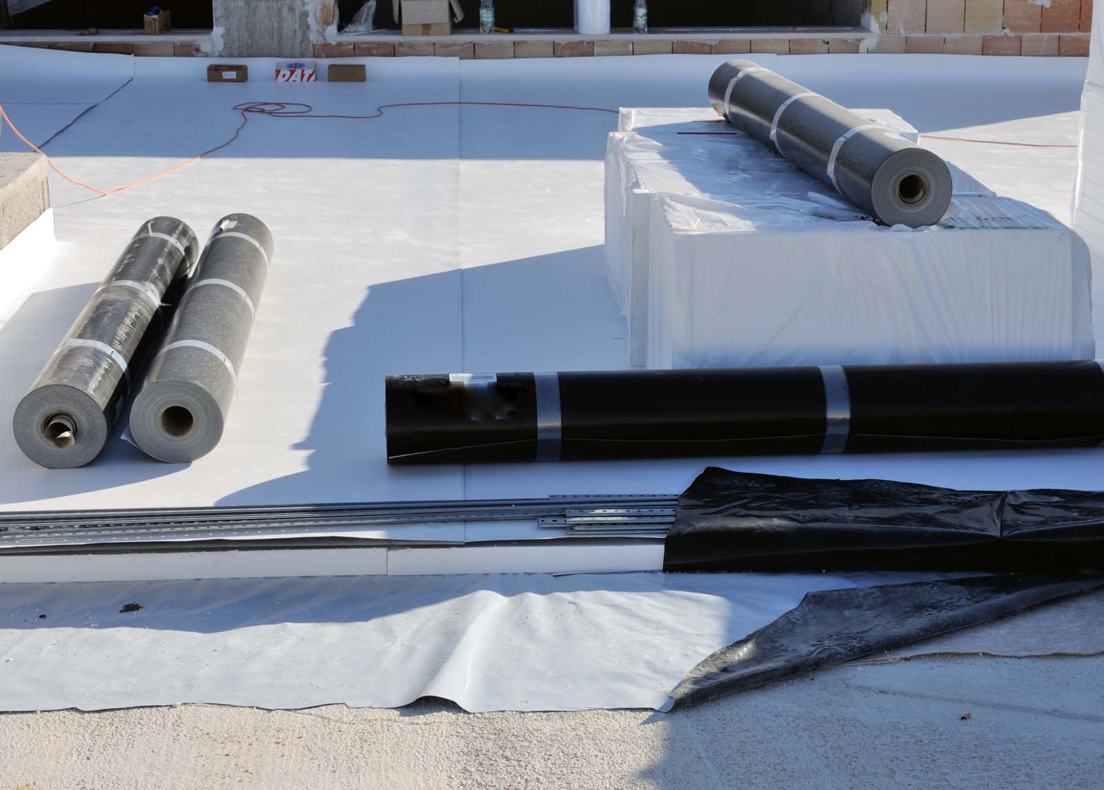 Several rolled roofing materials and tools, accompanied by options for metal roofing services, are placed on a flat surface, likely prepared for installation on a construction site.