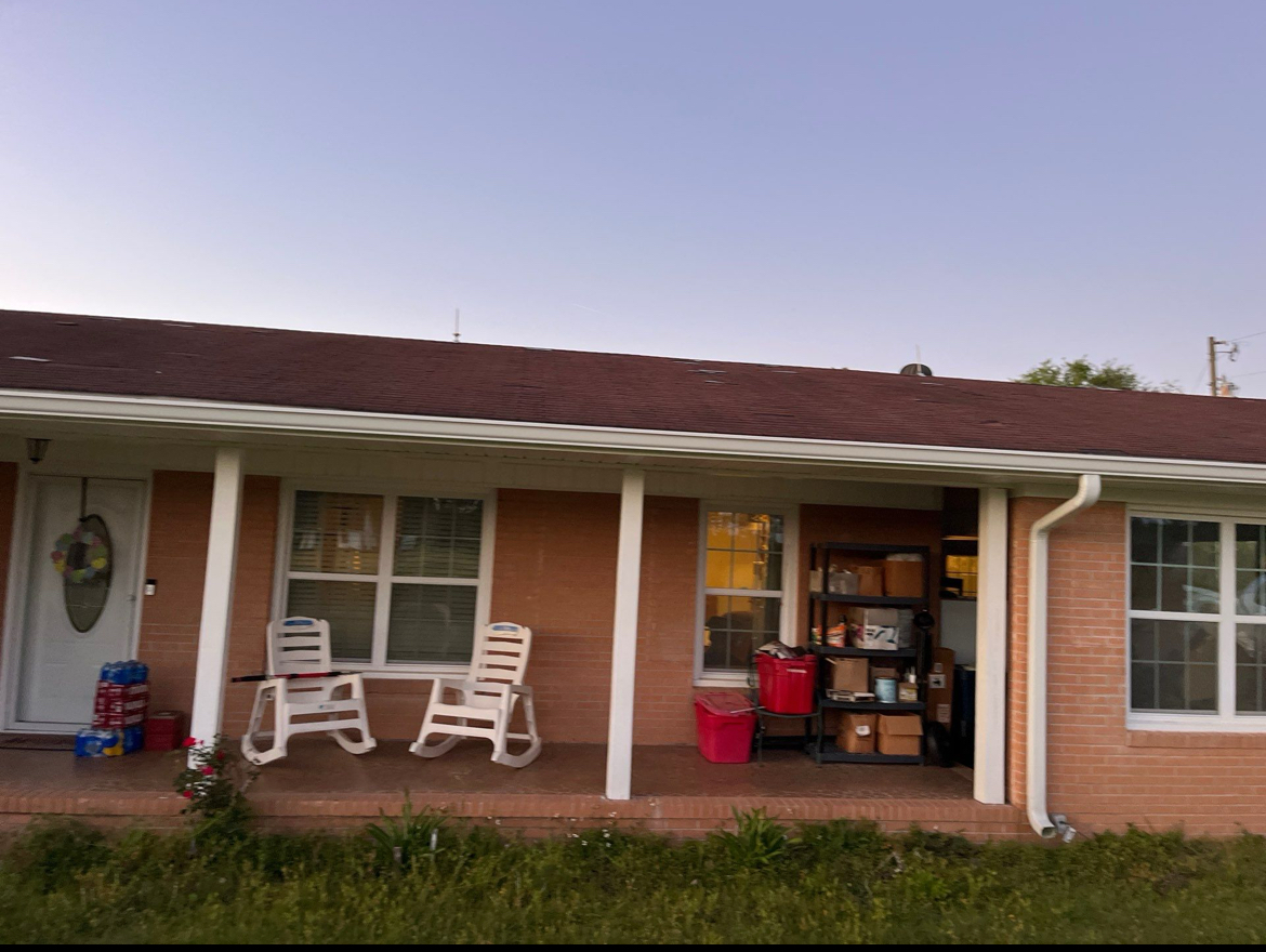 Single-story brick house with a porch featuring two white chairs and a small table. The porch also has stacked boxes and containers. The sky is clear.