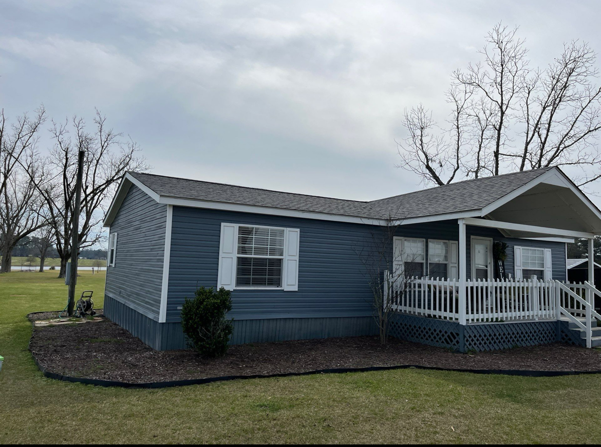 Single-story blue house with a white porch and railing. Surrounded by grass and bare trees. Overcast sky above.