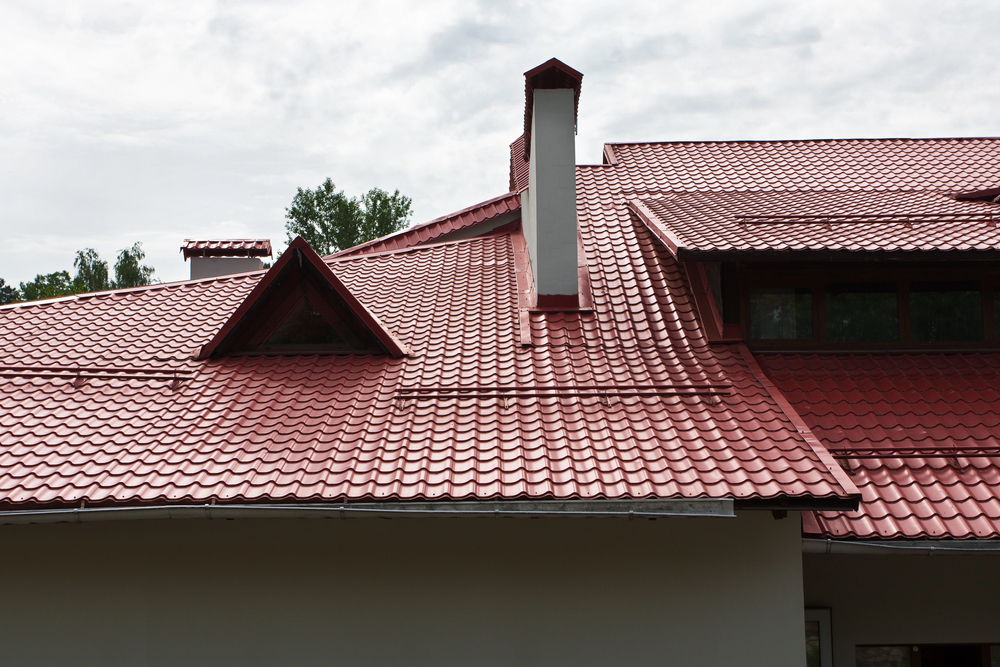 Red metal tiled roof with multiple peaks and a chimney against a cloudy sky.