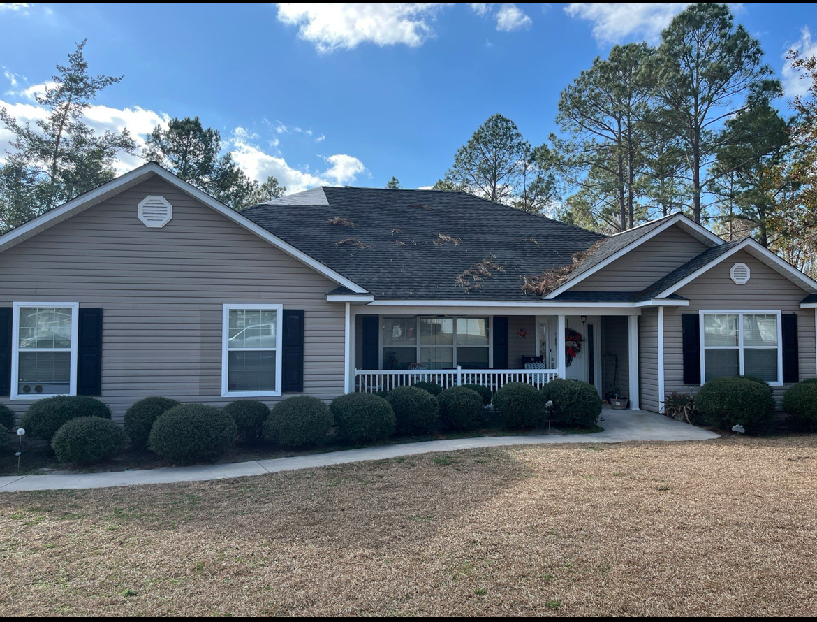 A single-story gray house with black shutters, a covered porch, and a front yard with shrubs. Trees and a partly cloudy sky are in the background.