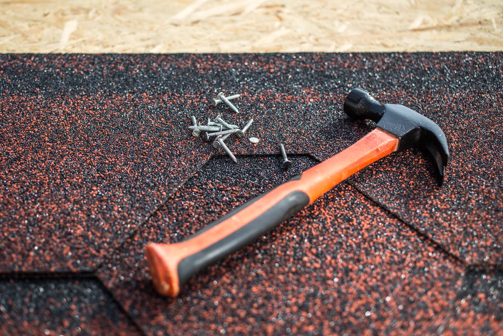 A hammer and several nails lie on red and black asphalt shingles, expertly positioned over a wooden surface, showcasing the craftsmanship of metal roofing services.