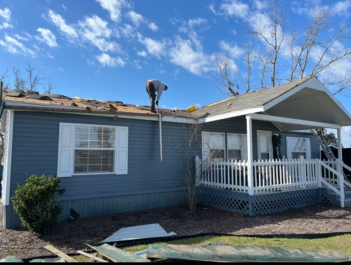 Person repairing a roof on a blue house with white trim and a porch, under a partly cloudy sky.